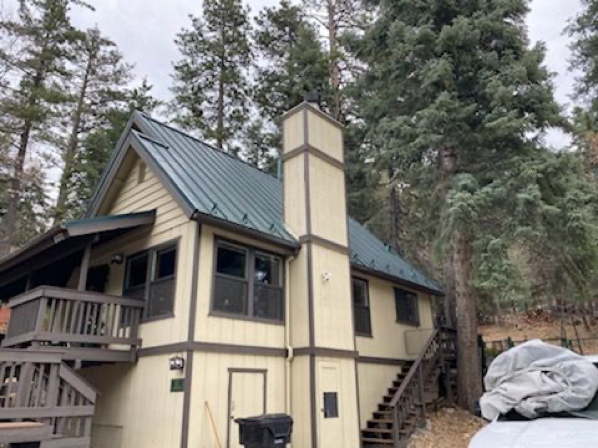 A mountain cabin under tall pines with a dark green standing seam metal roof and a board-and-batten chimney chase.