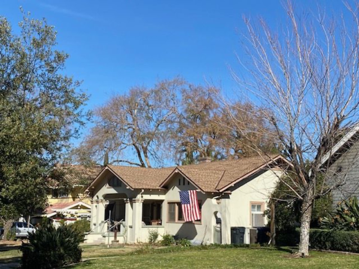 A craftsman bungalow with a new shingled roof and an American flag hanging from the porch, bare trees against a blue sky.