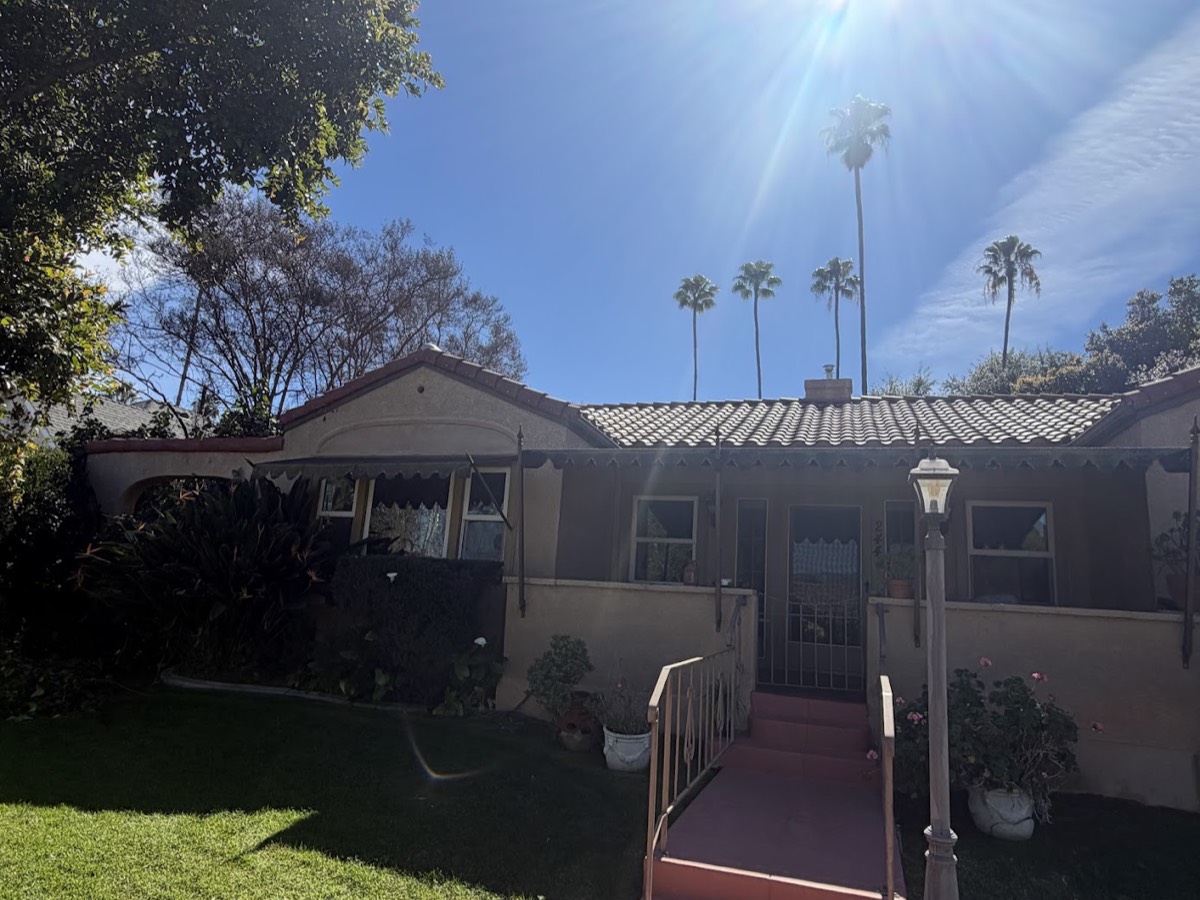 A single-story home with a light tile roof and palm trees behind it, sun flaring over the ridge.
