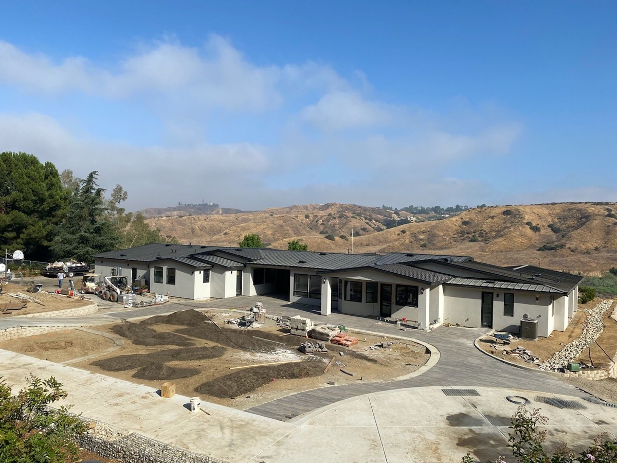 A long single-story house under construction on a hillside lot, with a dark roof already finished and graded dirt in front.